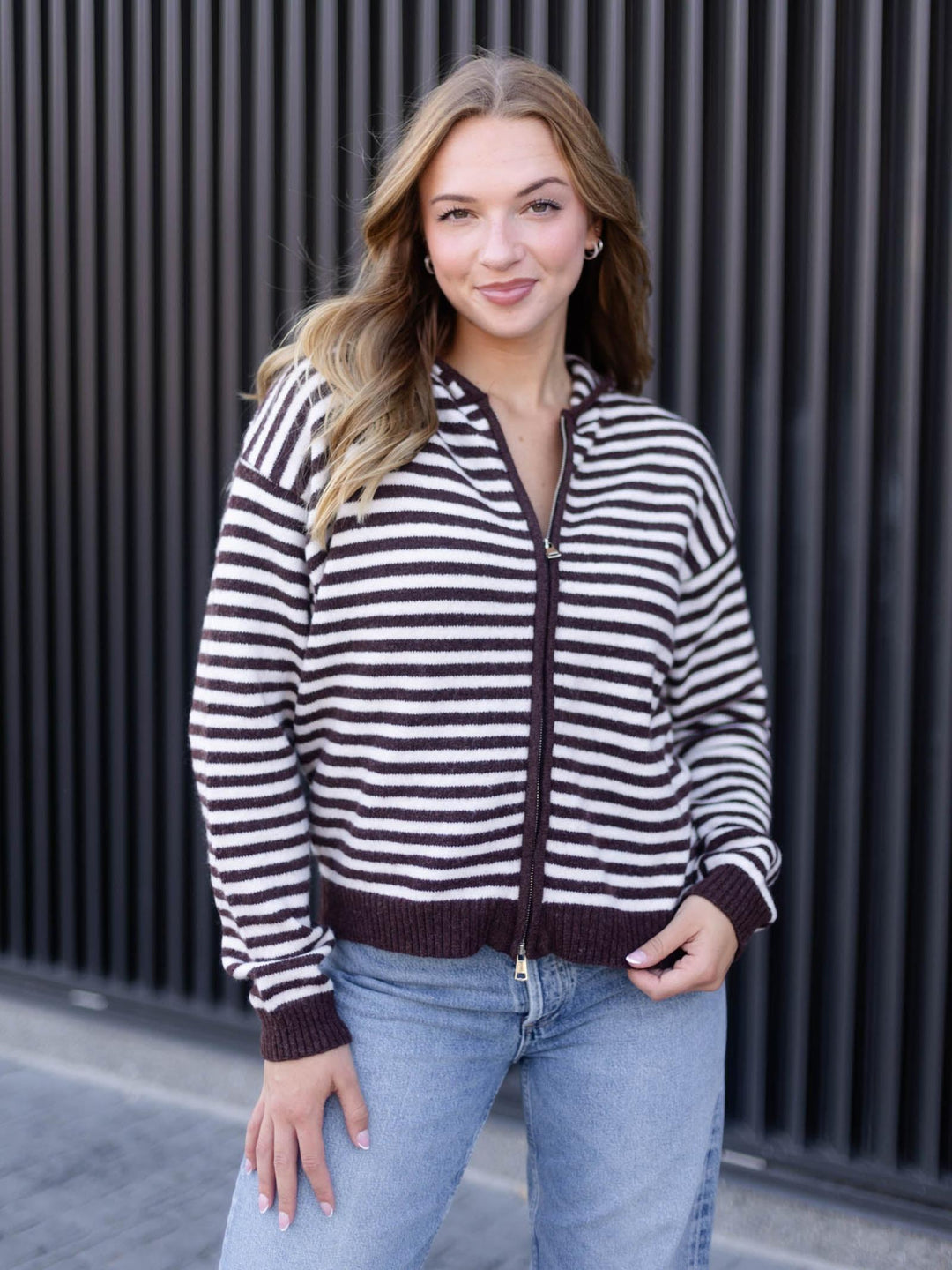 A woman wearing a brown and white horizontally striped zip-up knit cardigan with a hood, paired with light blue jeans, standing in front of a black slatted wall.