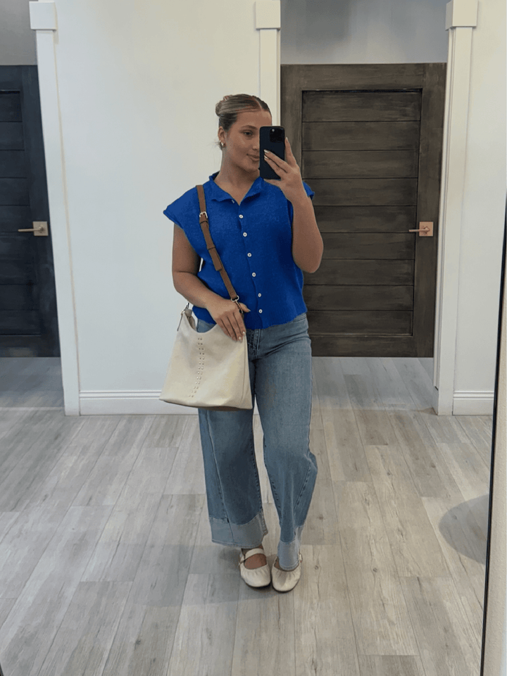Woman taking a mirror selfie wearing a blue top and jeans in a room with wooden flooring.