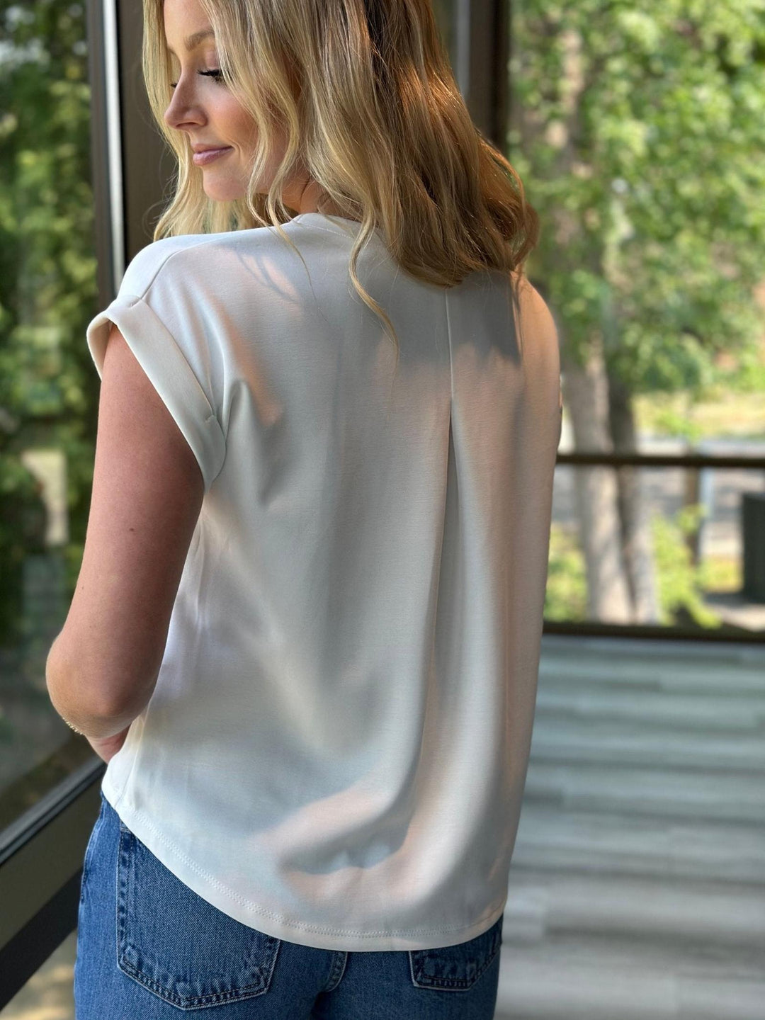 A woman wearing a short-sleeve, off-white top and blue jeans, photographed from behind in a bright indoor setting with a window and green outdoor scenery in the background.