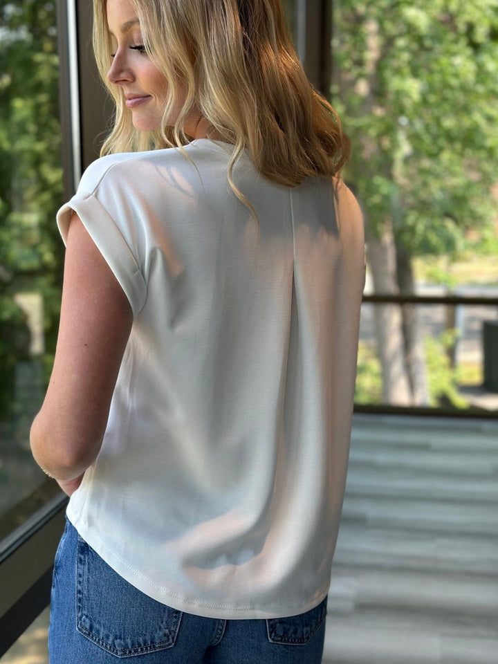A woman wearing a short-sleeve, off-white top and blue jeans, photographed from behind in a bright indoor setting with a window and green outdoor scenery in the background.