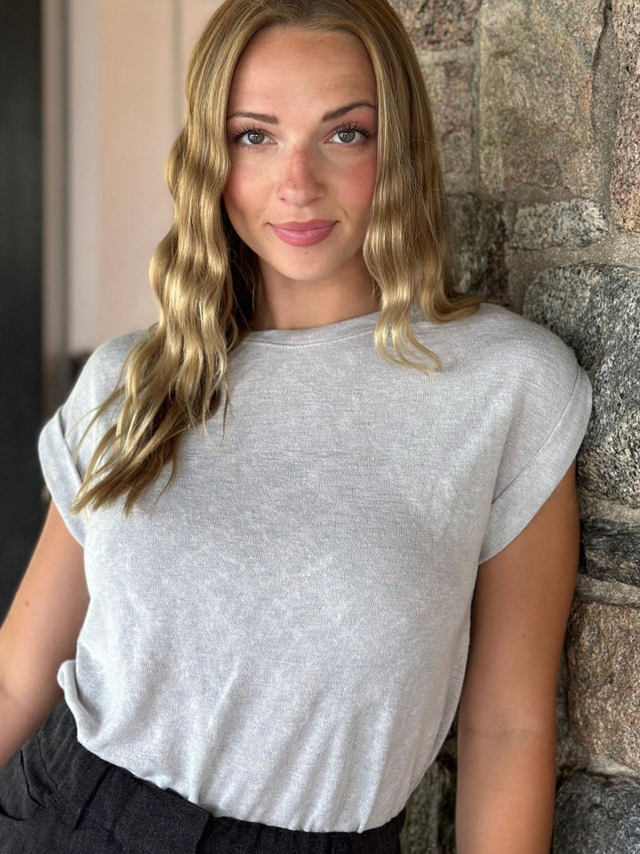 A woman with blonde wavy hair wearing a light gray heathered short-sleeve t-shirt and dark gray pants, leaning against a rustic stone wall.