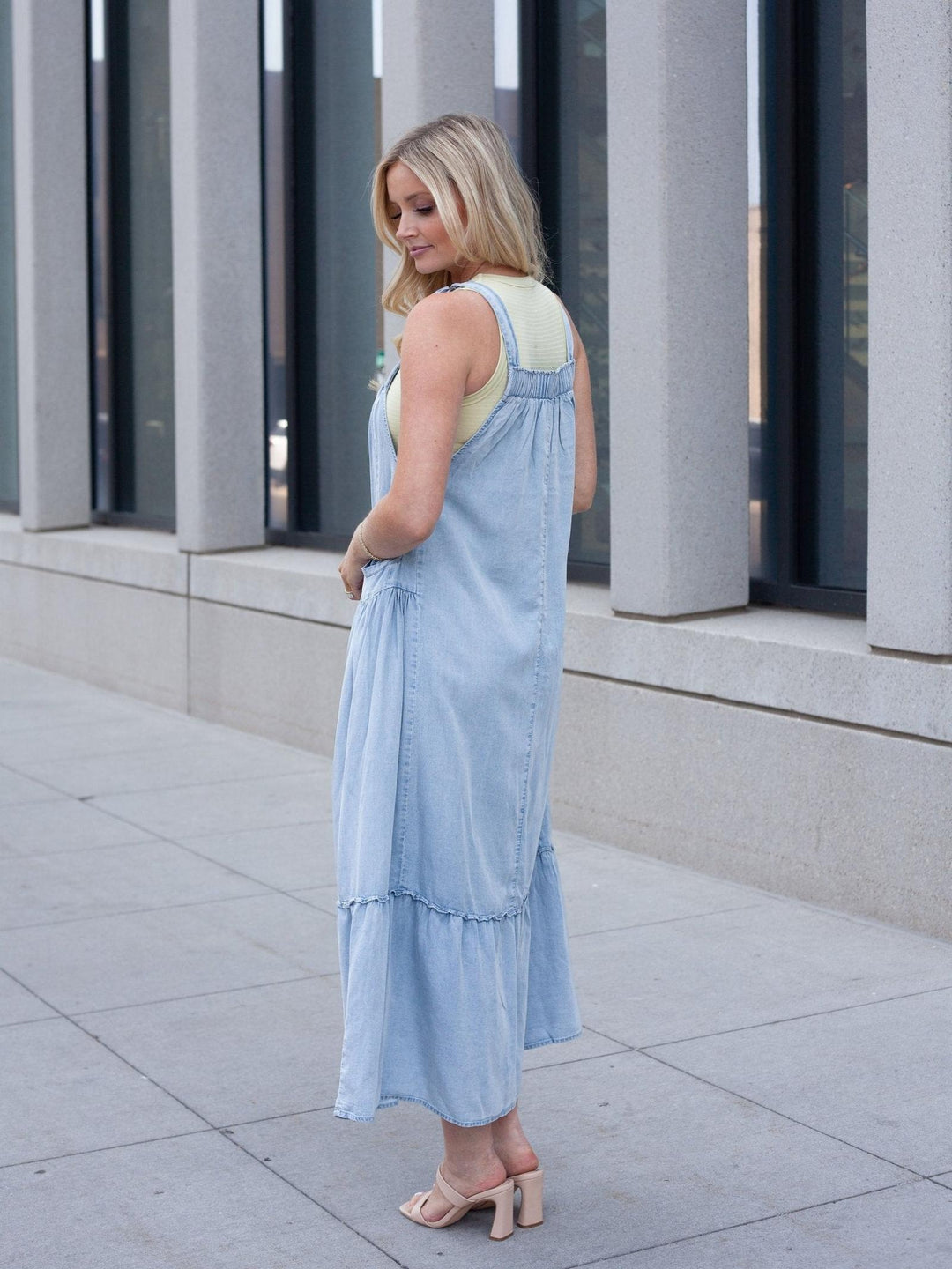 A woman wearing a light blue denim overall-style maxi dress over a pale yellow top, standing outdoors on a paved walkway in front of a modern building with large windows and columns.
