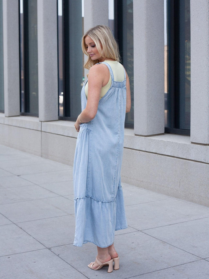 A woman wearing a light blue denim overall-style maxi dress over a pale yellow top, standing outdoors on a paved walkway in front of a modern building with large windows and columns.