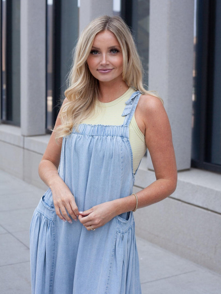 A blonde woman wearing a light blue denim overall dress over a pale yellow ribbed tank top, standing outdoors in front of a modern building with gray columns.