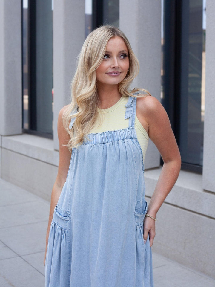 A blonde woman wearing a light blue denim overall dress over a pale yellow tank top, standing outdoors in front of a modern building with gray columns.
