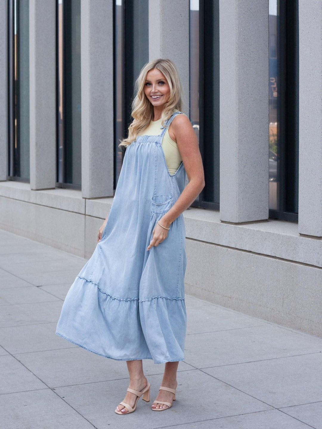A woman wearing a light blue denim overall midi dress over a pale yellow top, standing outdoors on a paved walkway in front of a modern building with gray columns.