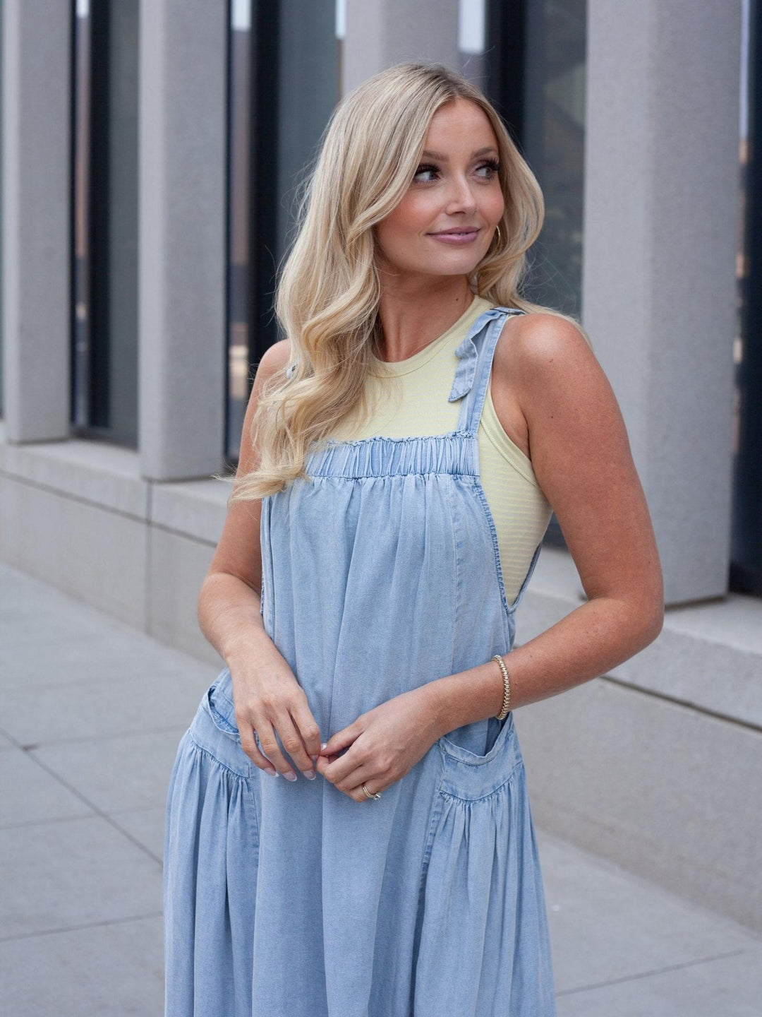 A blonde woman wearing a light blue denim overall dress over a pale yellow ribbed tank top, standing outdoors in front of a modern gray building.