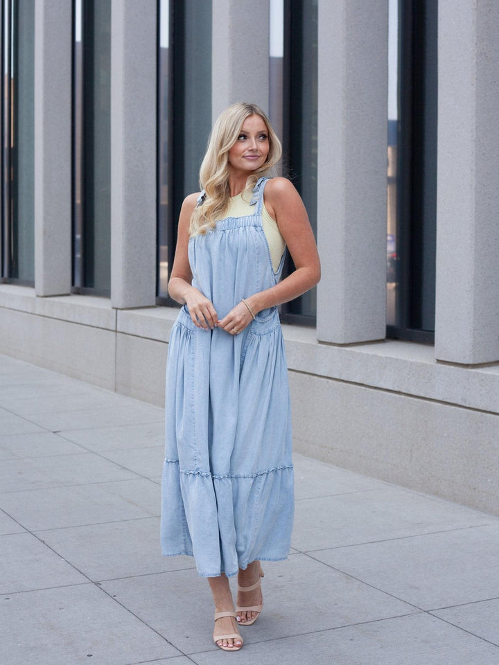 A woman wearing a light blue denim overall-style maxi dress with a pale yellow tank top underneath, standing on a paved walkway in front of a modern building with gray columns.