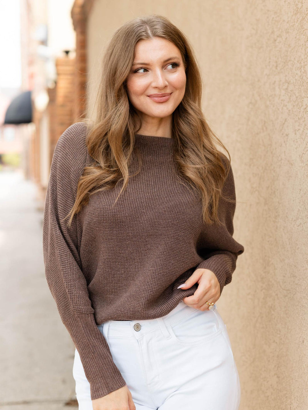 A woman with long light brown hair wearing a brown ribbed knit sweater and white pants, standing outdoors against a beige stucco wall.