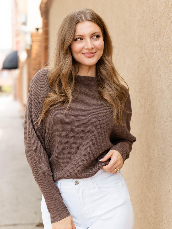 A woman with long light brown hair wearing a brown ribbed knit sweater and white pants, standing outdoors against a beige stucco wall.