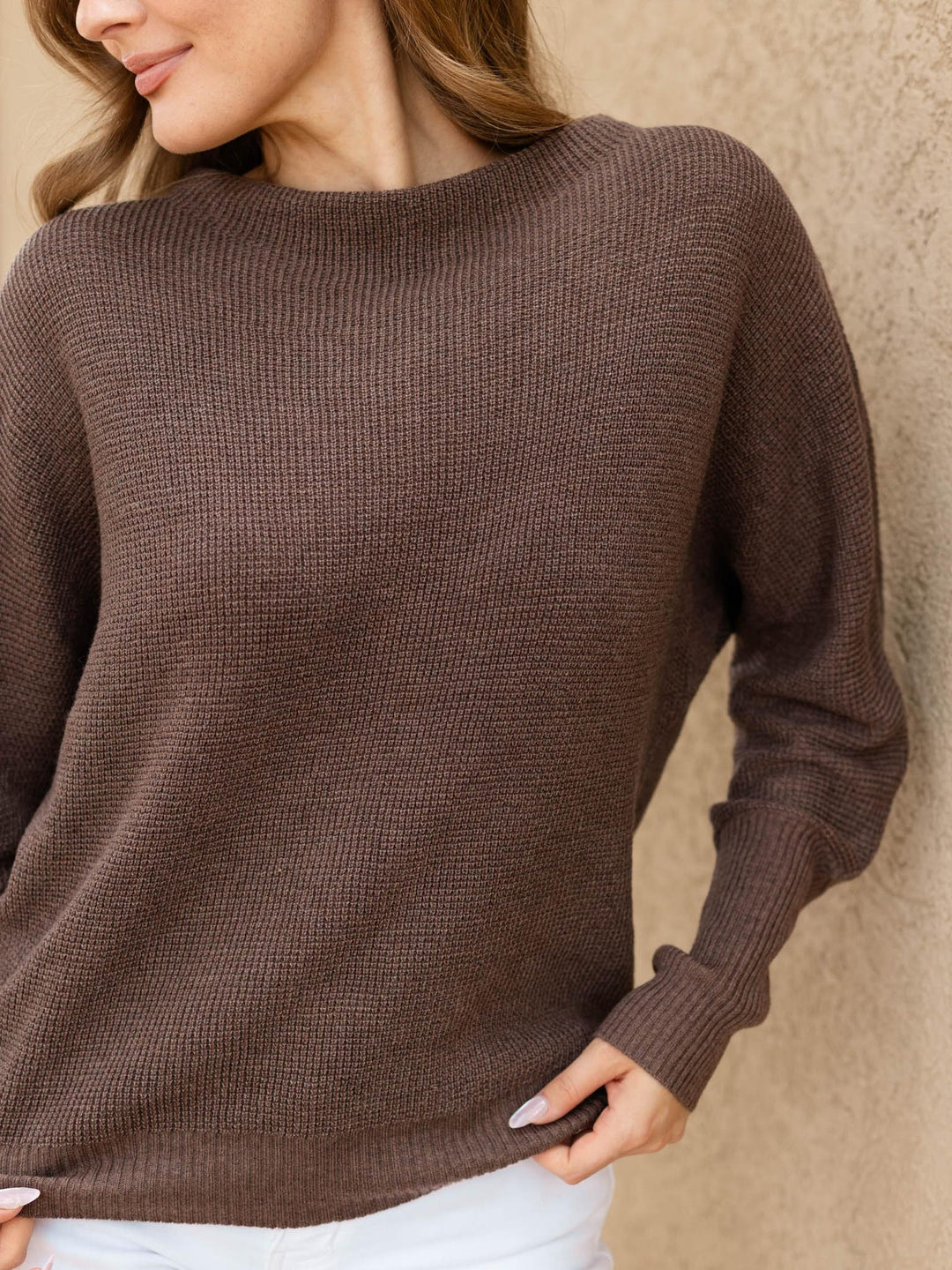 A woman wearing a brown waffle-knit long-sleeve sweater, shown from the side against a textured beige wall.