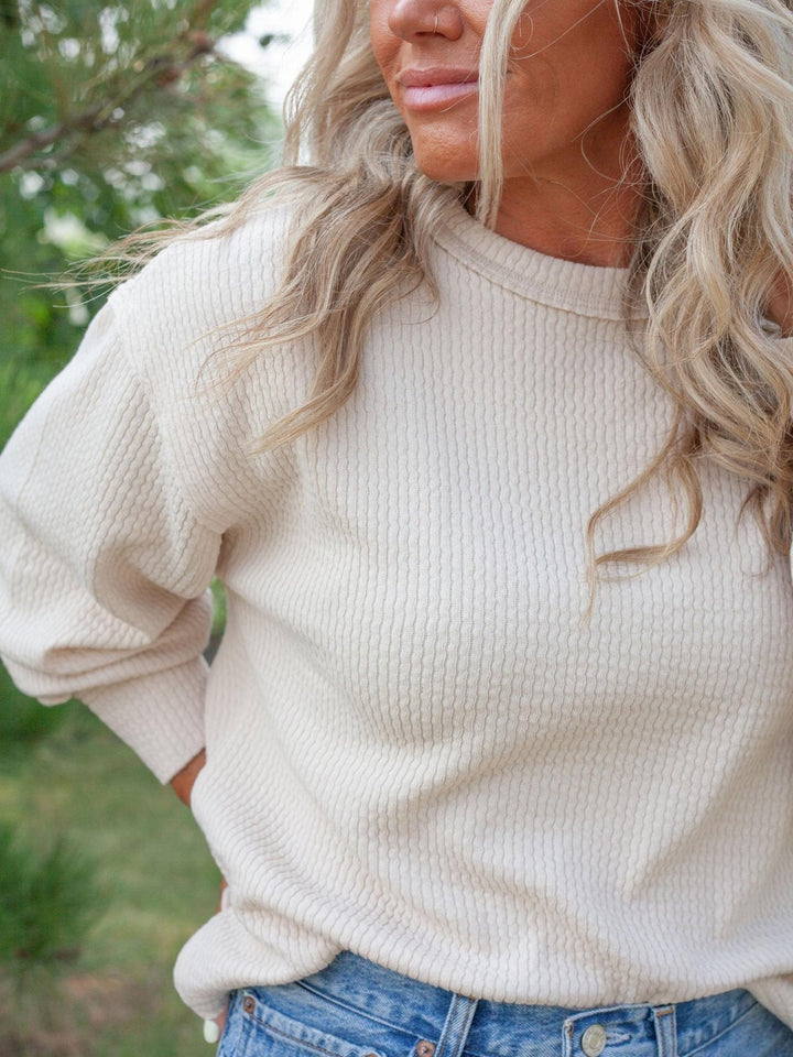A woman wearing a cream-colored waffle-knit crewneck sweater, paired with blue jeans, photographed outdoors with a soft-focus green background.