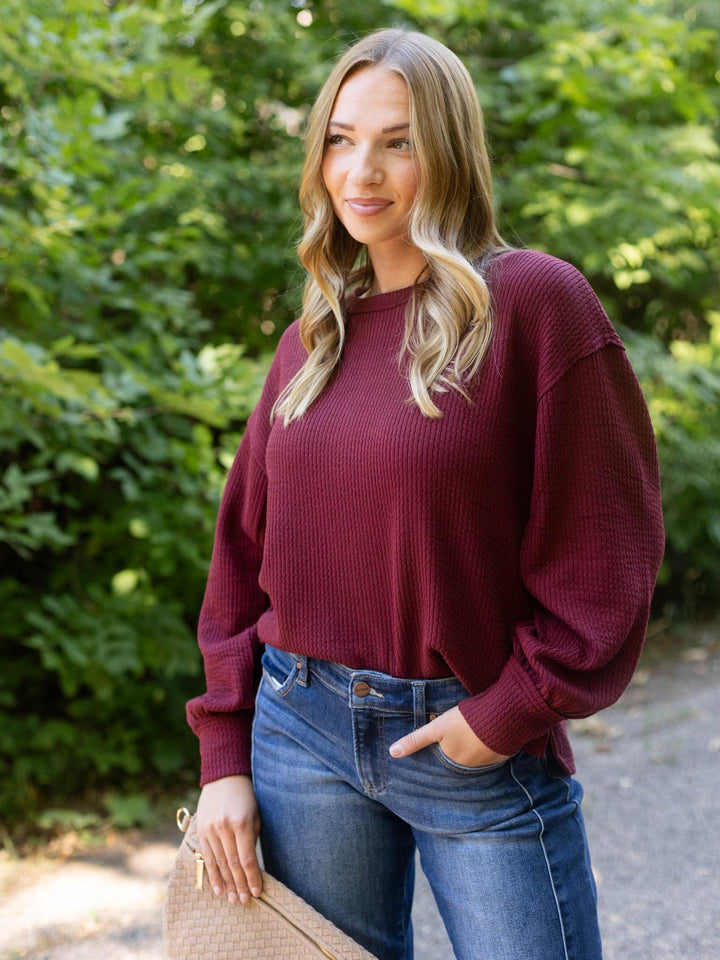 A woman wearing a maroon waffle-knit long-sleeve sweater and blue jeans, standing outdoors in front of a green leafy background.