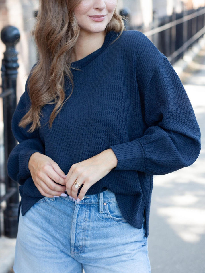A woman wearing a dark navy blue waffle-knit sweater and light blue jeans, standing outdoors on a city sidewalk.