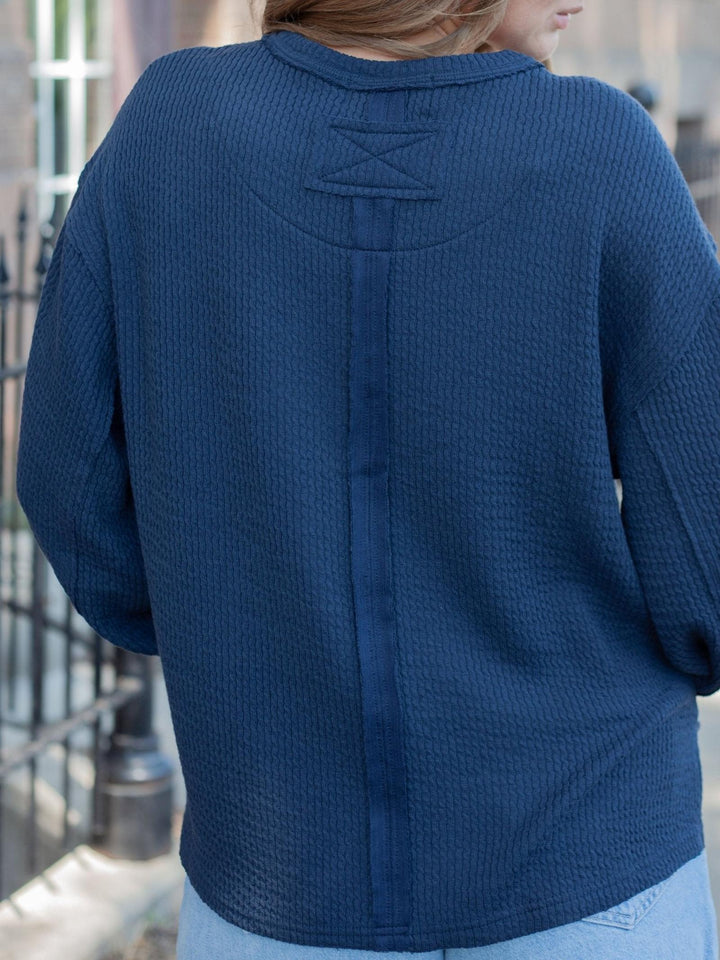 A person viewed from the back wearing a navy blue textured knit cardigan or sweater with a decorative stitched X detail across the upper back.