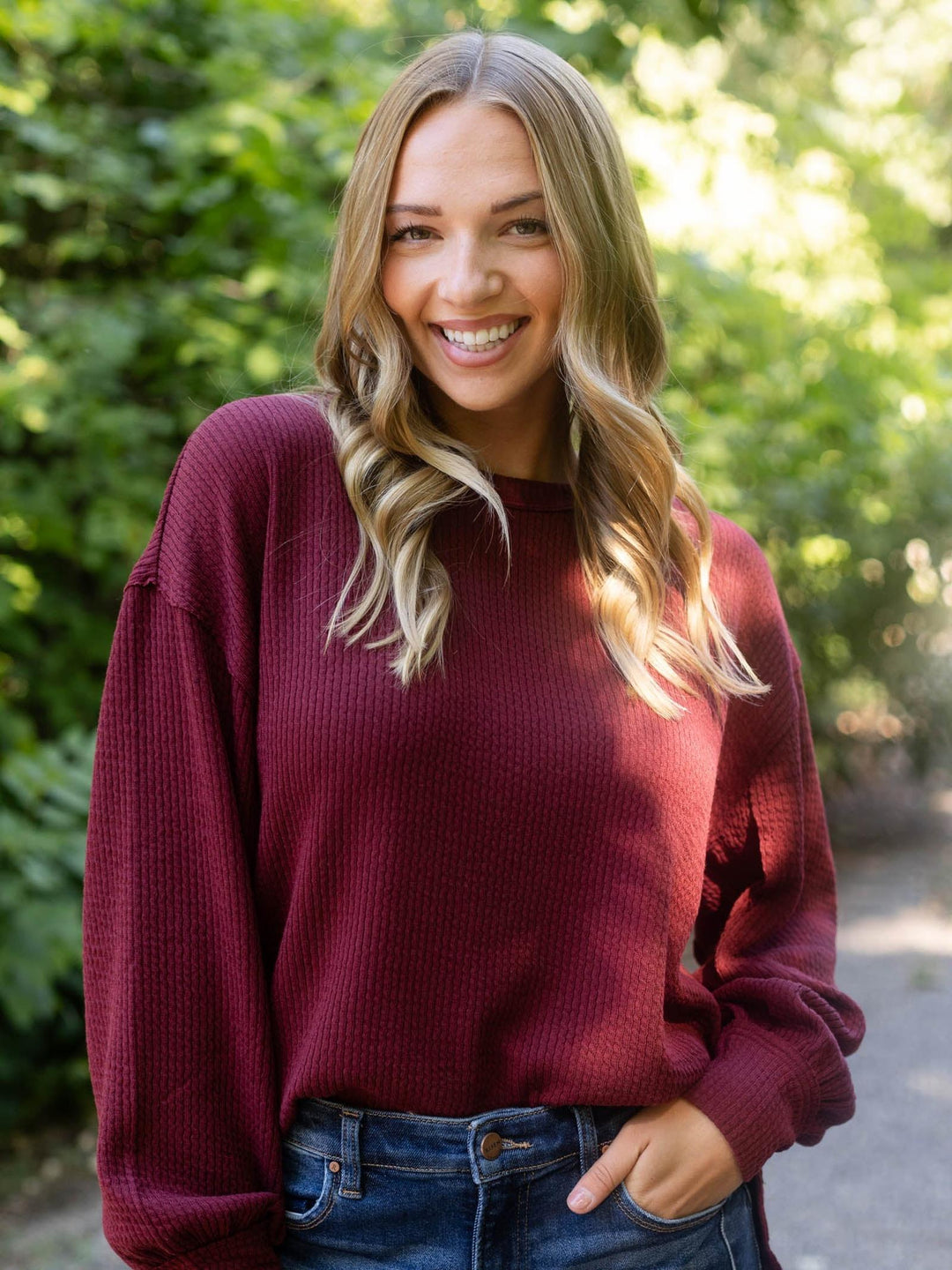 A smiling woman with blonde hair wearing a maroon waffle-knit long-sleeve sweater and blue jeans, standing outdoors against a blurred green foliage background.