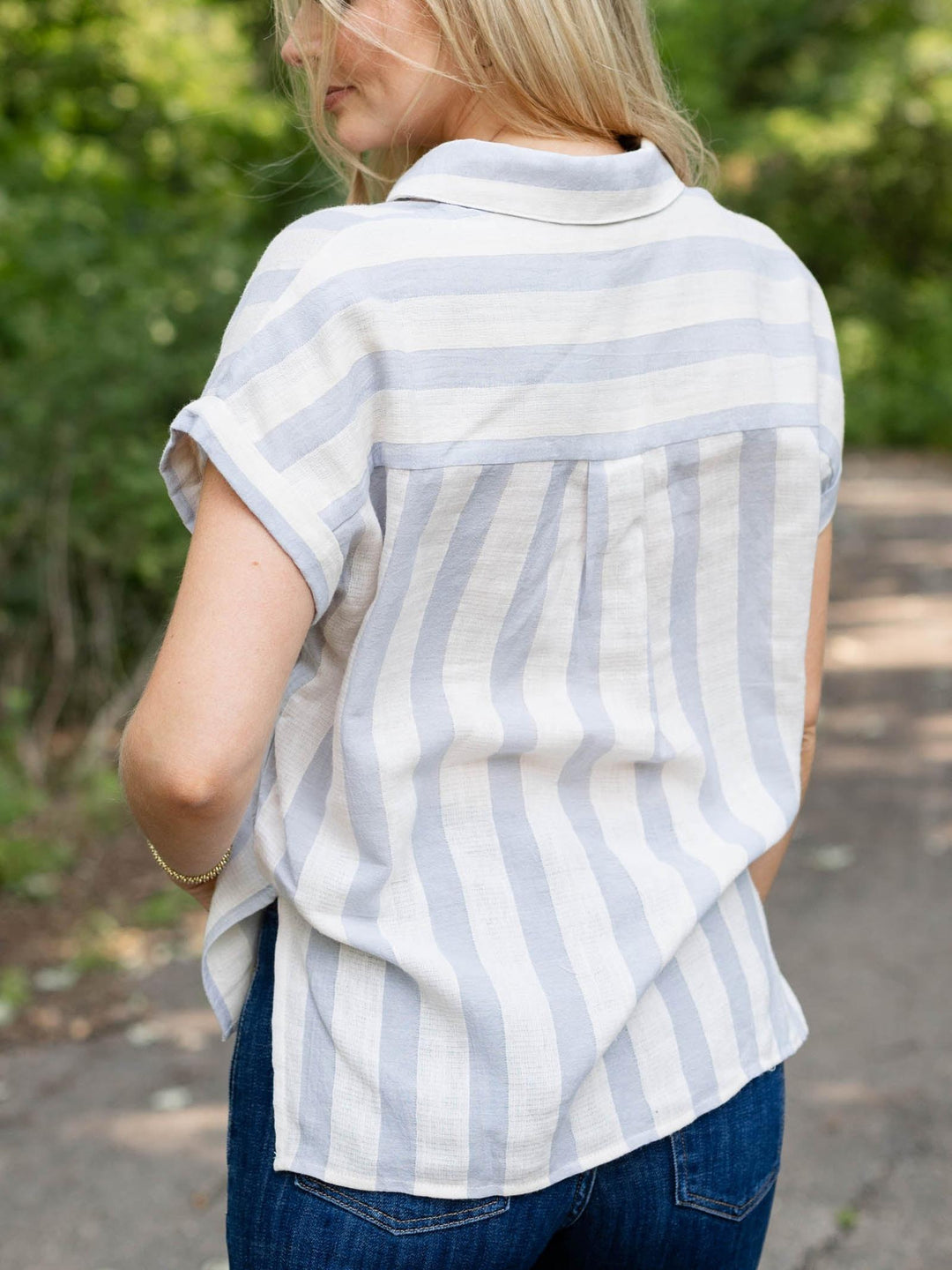 A woman viewed from behind wearing a short-sleeve collared shirt with vertical light blue and white stripes, paired with blue jeans, outdoors on a path with green foliage in the background.