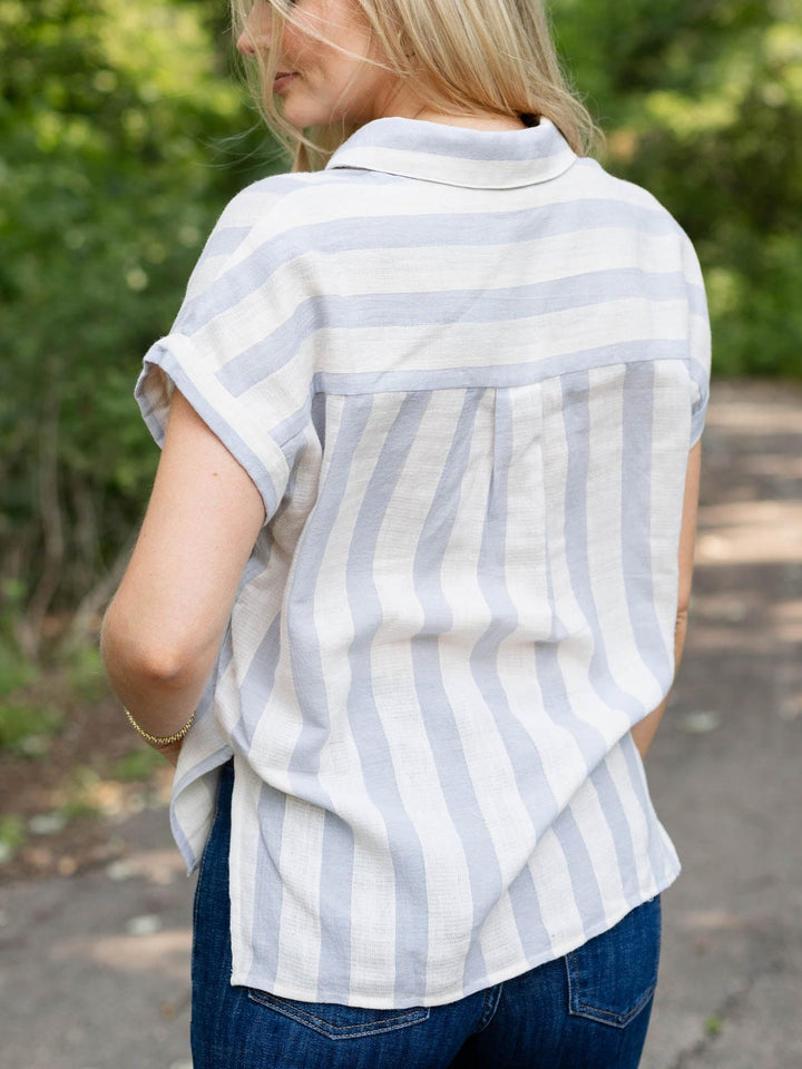 A woman viewed from behind wearing a short-sleeve collared shirt with vertical light blue and white stripes, paired with blue jeans, outdoors on a path with green foliage in the background.