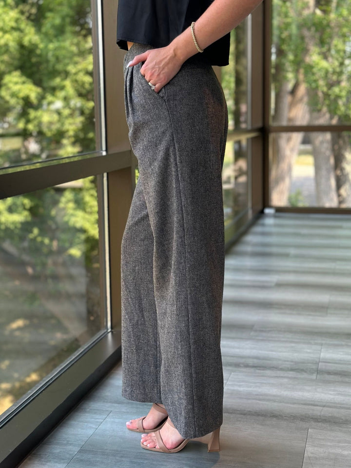 A person wearing high-waisted, wide-leg gray trousers with a textured heathered finish, paired with a black top and light pink heeled sandals, standing in a modern indoor space with large windows overlooking green trees.
