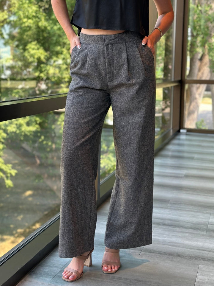 A person wearing high-waisted, wide-leg gray textured trousers with a black sleeveless top and beige heeled sandals, standing on a balcony with a view of green trees.