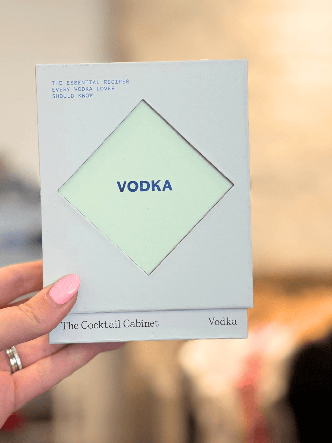 A hand with pink nail polish holds a square white card or booklet featuring a pale green diamond shape in the center with the word &