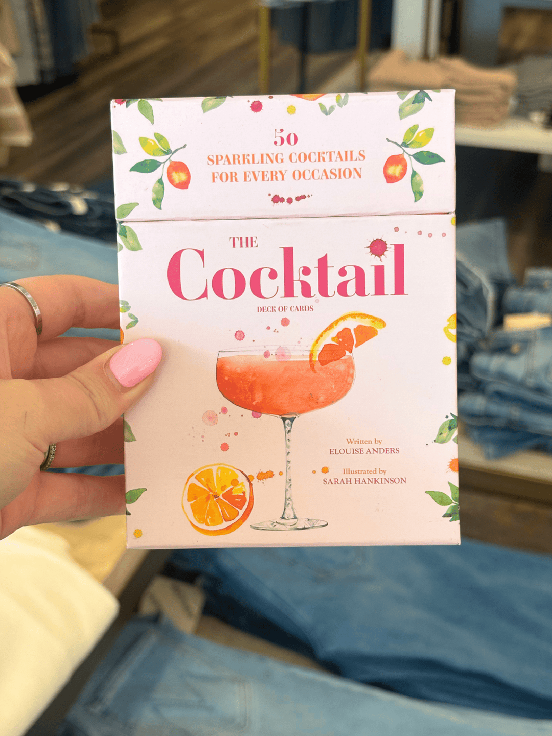 A hand with pink nail polish holds a book titled "The Cocktail" in front of a blurred background of blue jeans and clothing racks. The book cover is light pink with illustrations of orange slices, green leaves, and a pink cocktail glass.