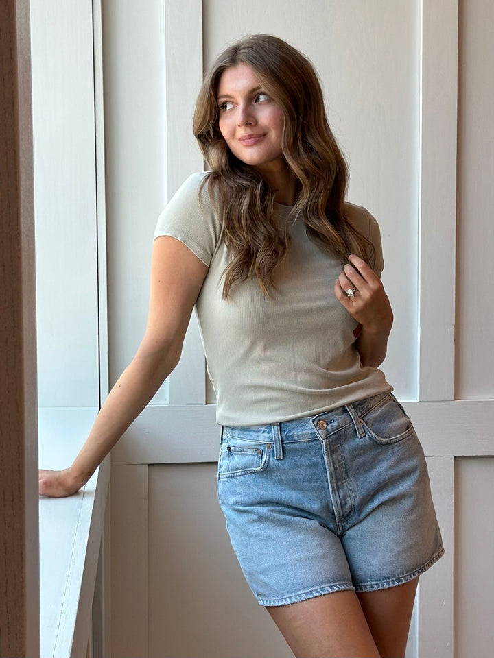 A woman with long brown hair stands indoors near a window, wearing a fitted short-sleeve beige t-shirt and light blue denim shorts.