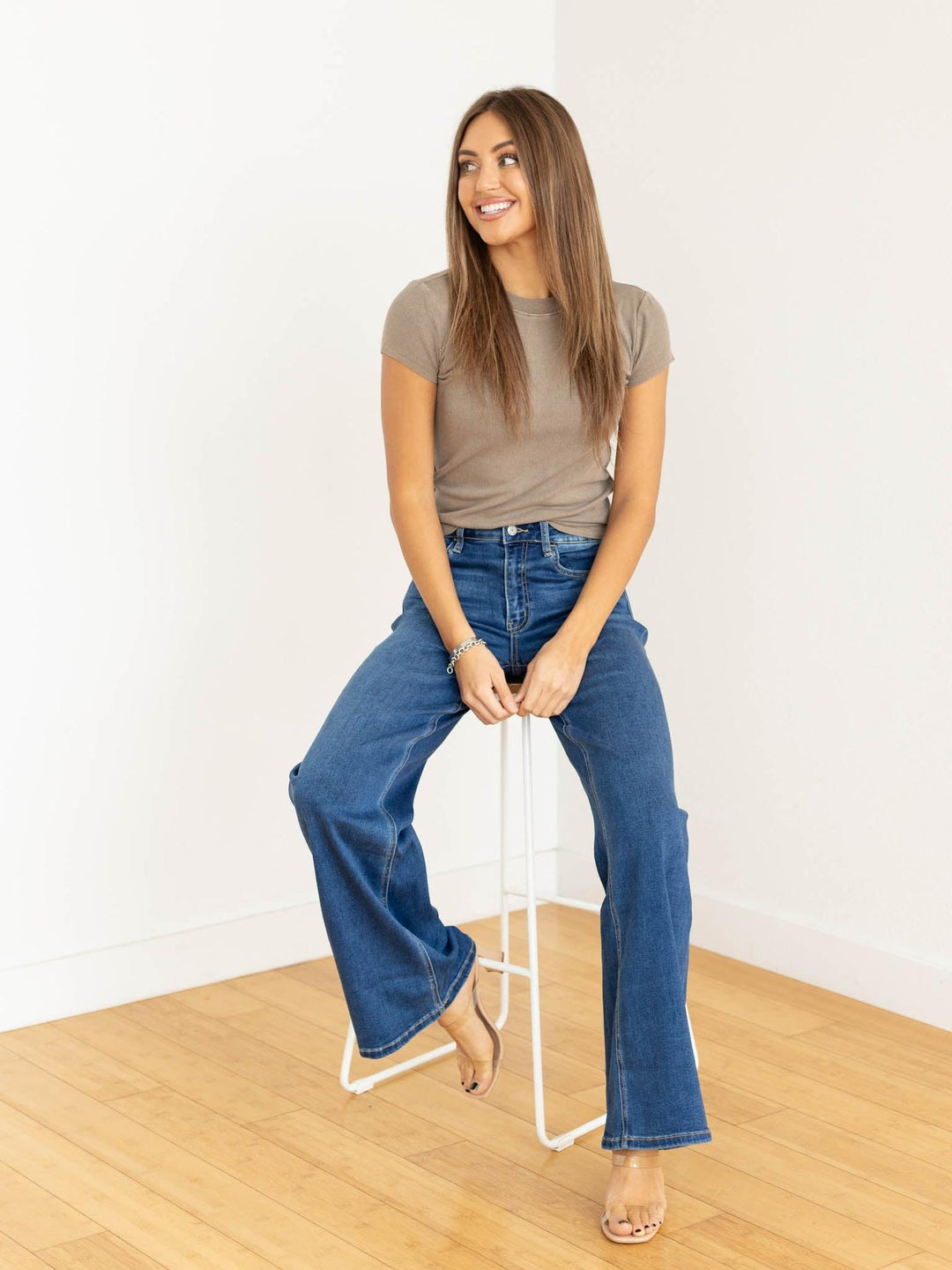 Woman sitting on a stool wearing a brown t-shirt and blue jeans against a white background