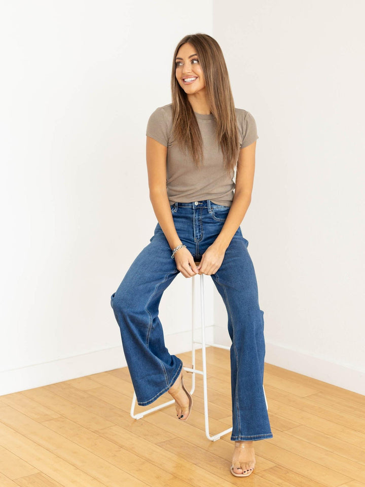 Woman sitting on a stool wearing a brown t-shirt and blue jeans against a white background