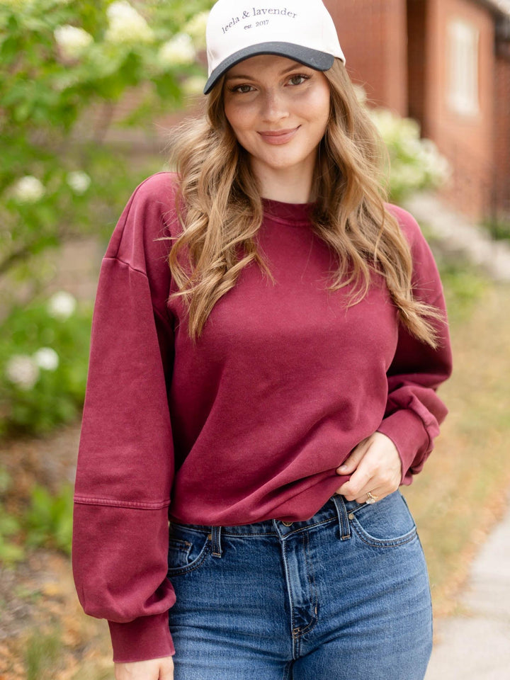 A woman wearing a washed burgundy crewneck sweatshirt and blue jeans, standing outdoors on a sidewalk with a brick building and greenery in the background. She is also wearing a white and navy baseball cap with &