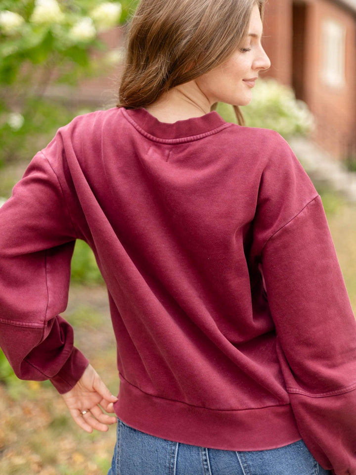 A woman wearing a dusty burgundy crewneck sweatshirt and blue jeans, photographed from the back in an outdoor setting with a blurred background.