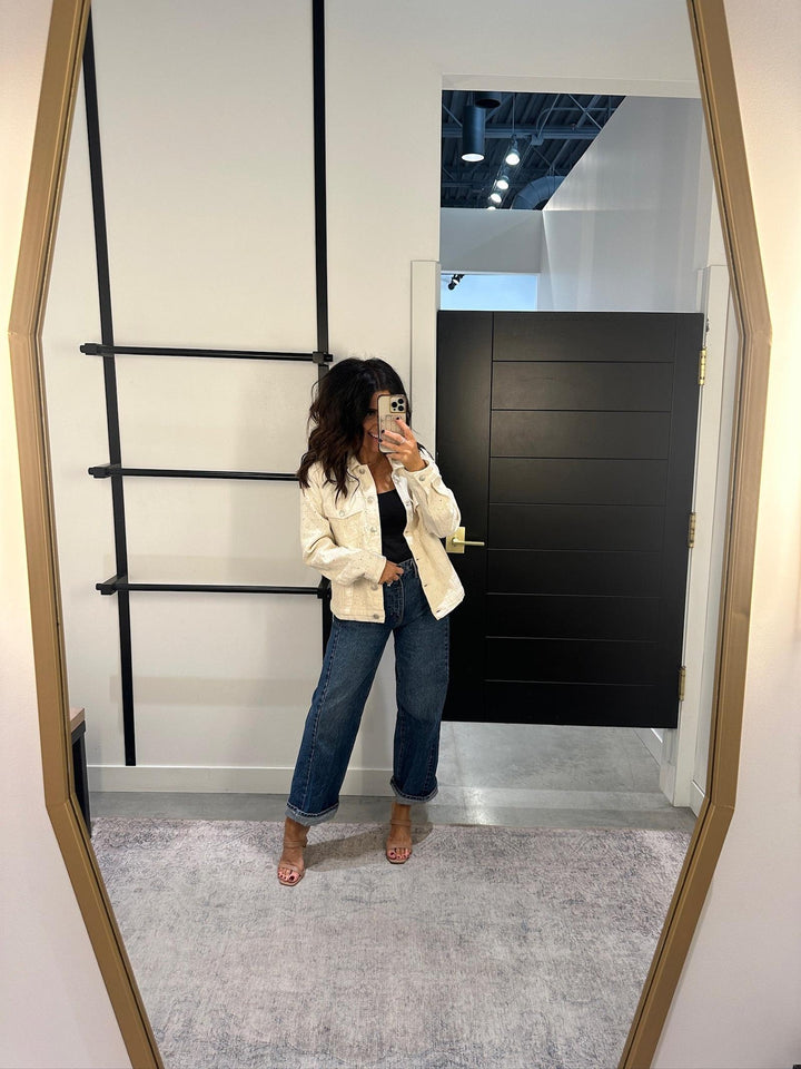 Person taking a mirror selfie wearing a white jacket and blue jeans in a modern interior setting.