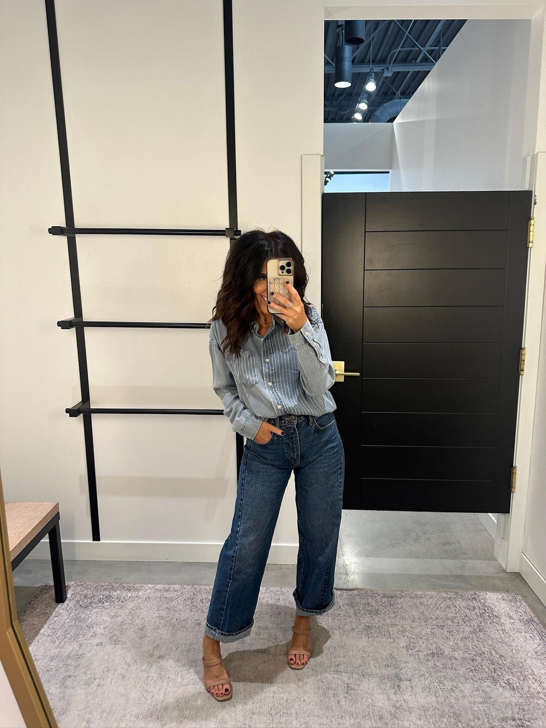 Person taking a mirror selfie wearing a checkered shirt and jeans in a room with a black door and ladder.