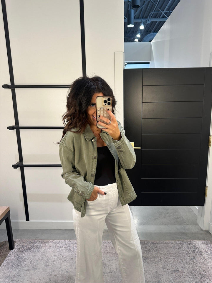 Woman taking a mirror selfie wearing an olive green jacket, black top, and white pants in a modern interior setting.