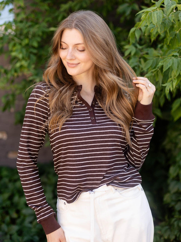 A woman with long wavy brown hair wearing a brown and white striped long-sleeve henley shirt and white pants, standing outdoors in front of green foliage.