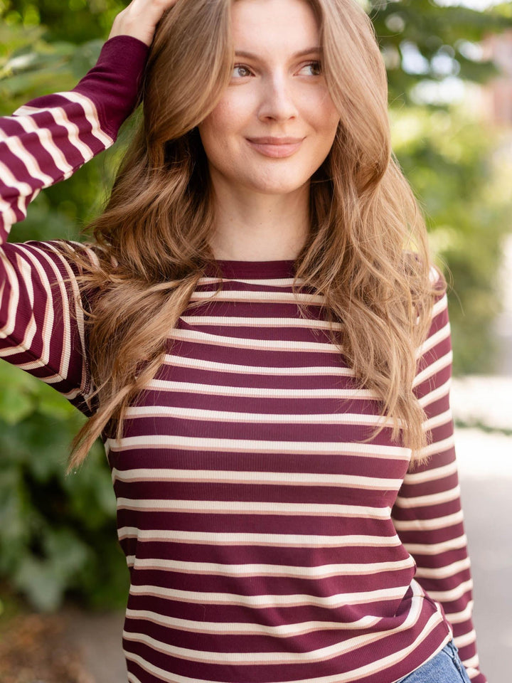 A woman wearing a long-sleeve striped knit top with burgundy and cream horizontal stripes, photographed outdoors with blurred green foliage in the background.
