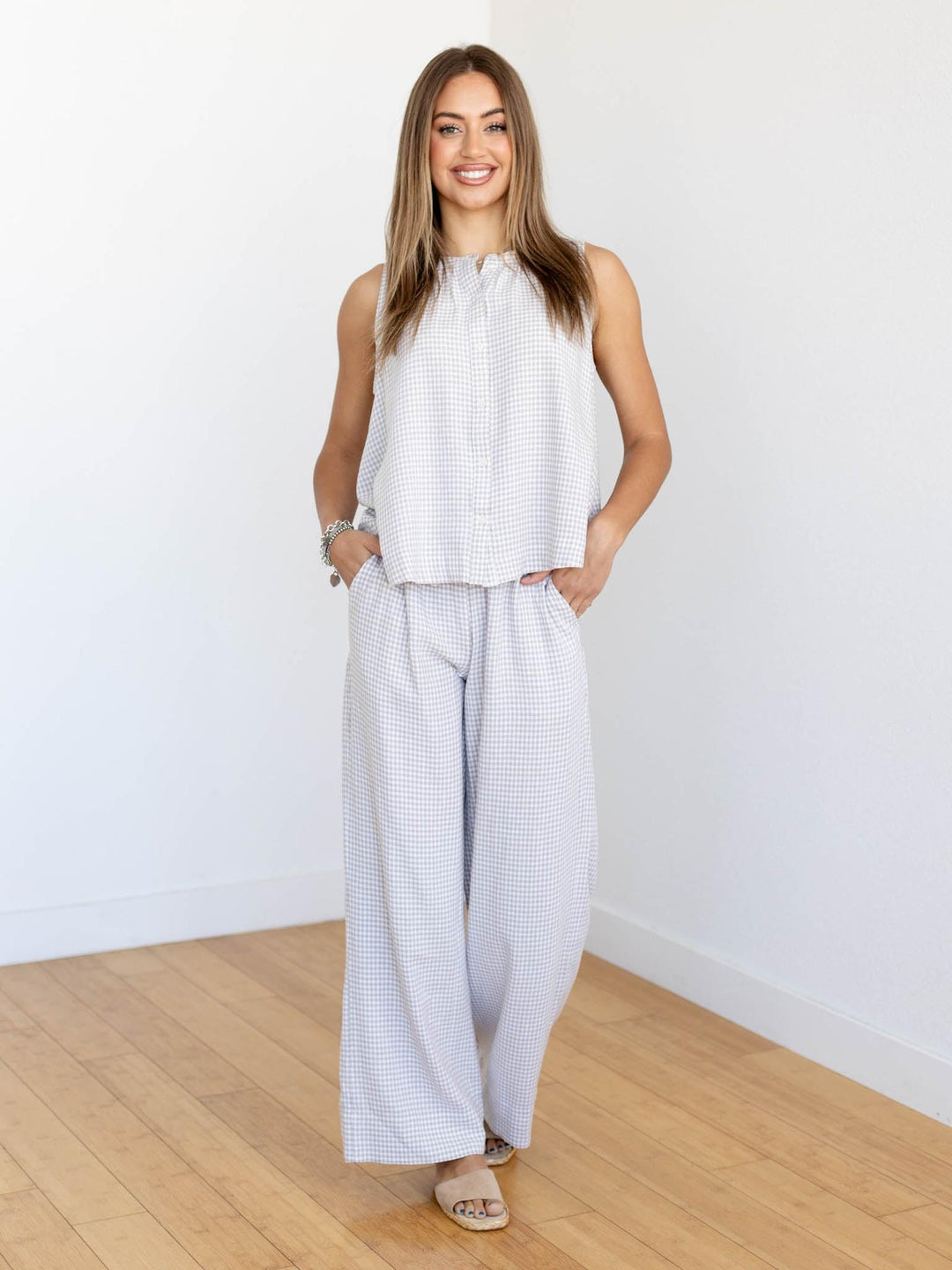 Woman wearing a white sleeveless top and wide-leg pants standing in a room with wooden flooring and white walls.