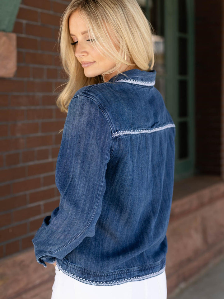 Woman wearing a blue denim jacket with white trim, standing in front of a brick wall.