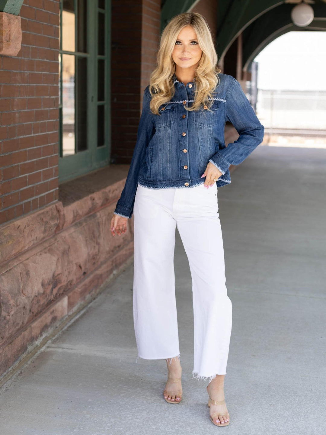 Woman wearing a denim jacket and white pants standing on a sidewalk.