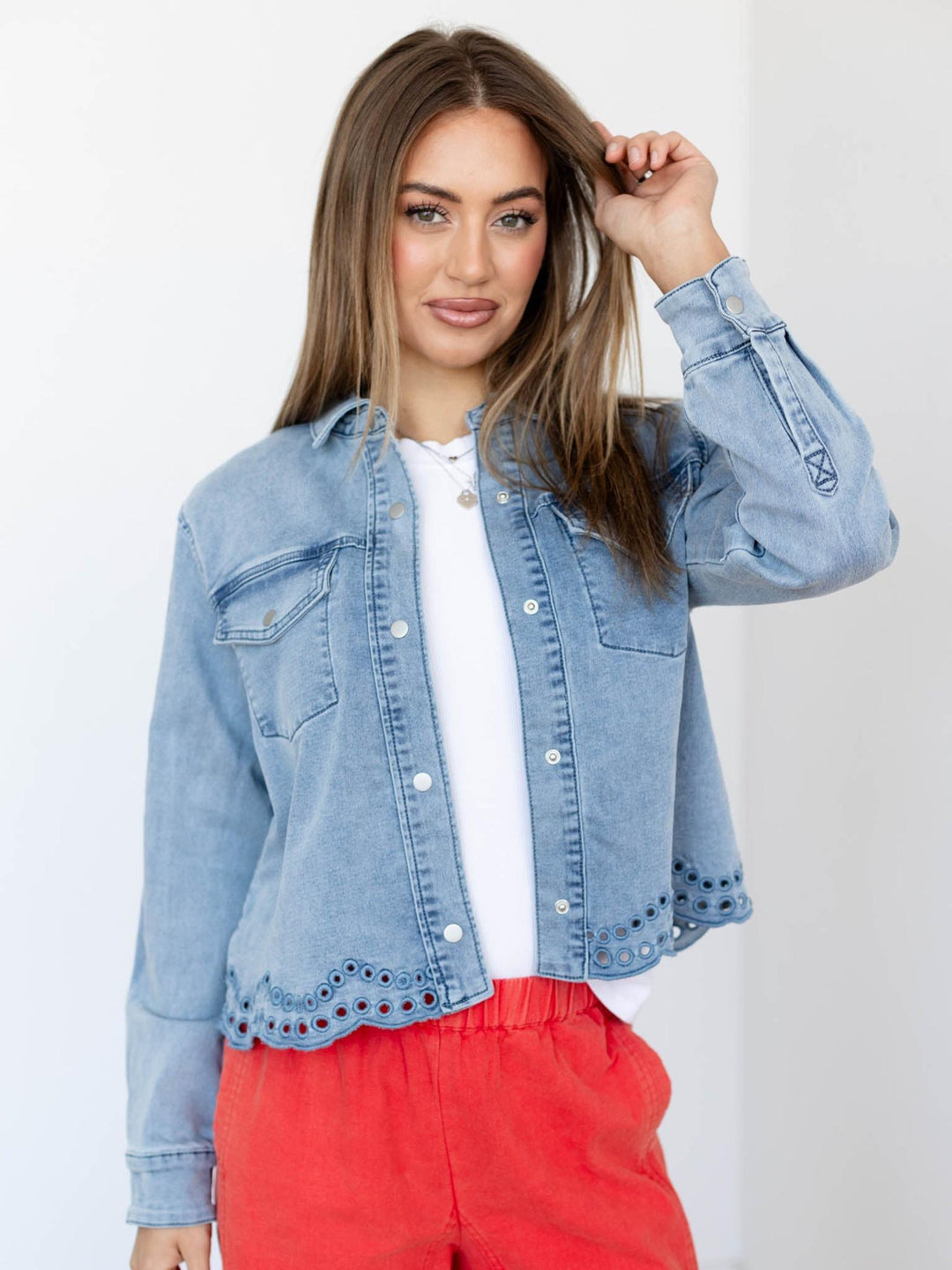 Woman wearing a light blue denim jacket over a white shirt with red pants on a plain background