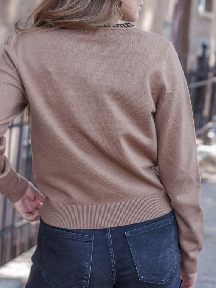 A woman viewed from behind wearing a camel brown knit sweater and dark blue jeans, standing outdoors on a city sidewalk.