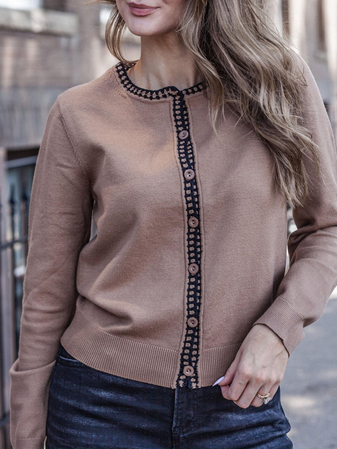 A woman wearing a tan knit cardigan with a dark patterned trim and button placket, paired with dark blue jeans.