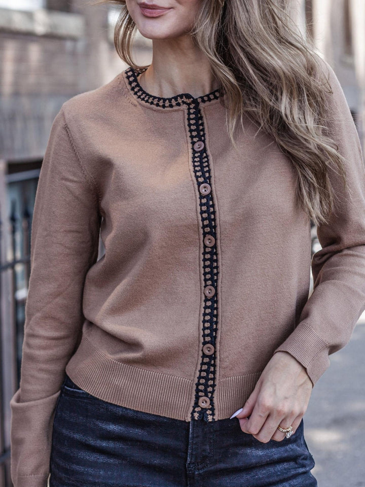 A woman wearing a tan knit cardigan with a dark patterned trim and button placket, paired with dark blue jeans.
