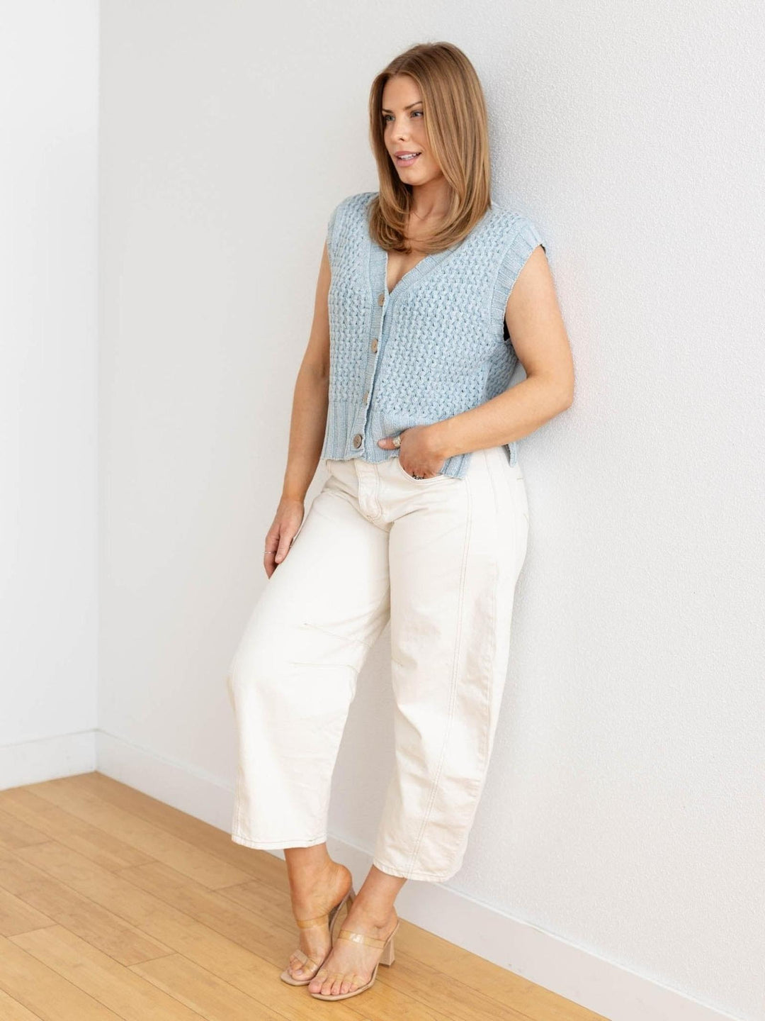 Woman wearing a light blue sleeveless top and white pants against a white wall.