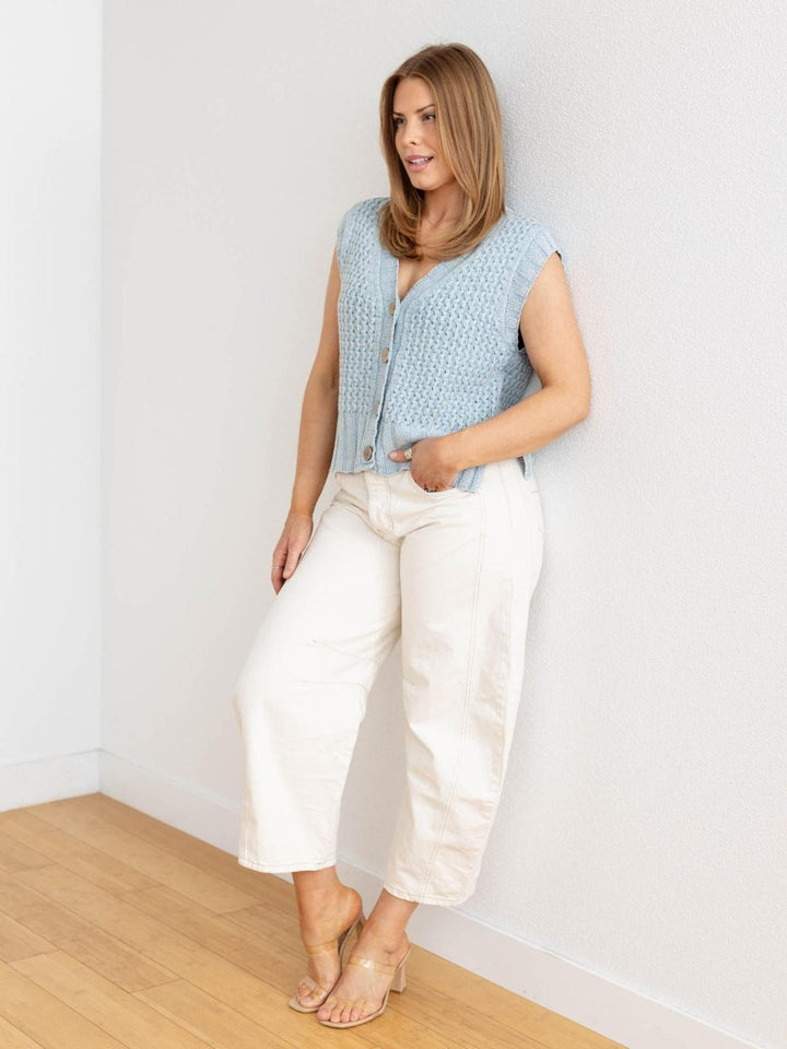 Woman wearing a light blue sleeveless top and white pants against a white wall.
