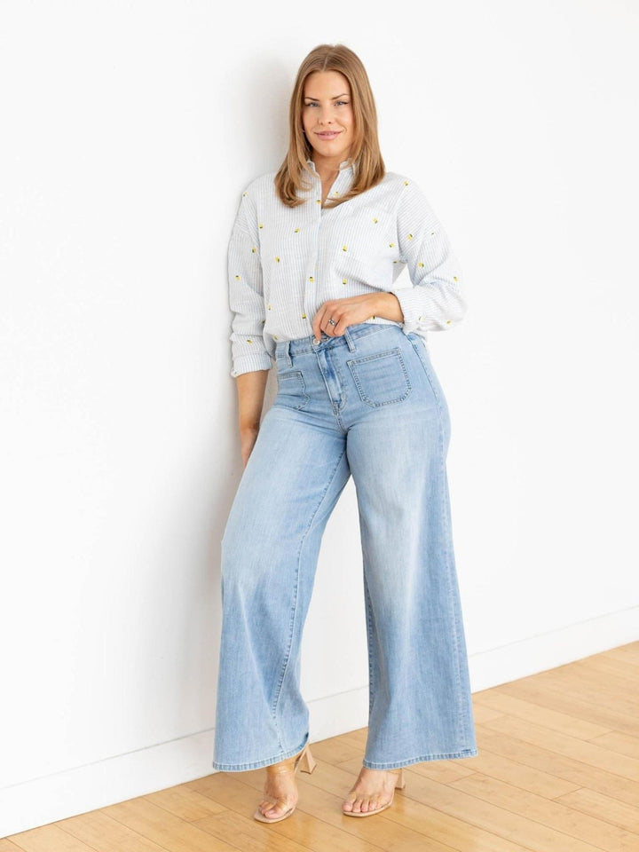 Woman wearing light blue jeans and a white blouse with gold buttons against a white wall.