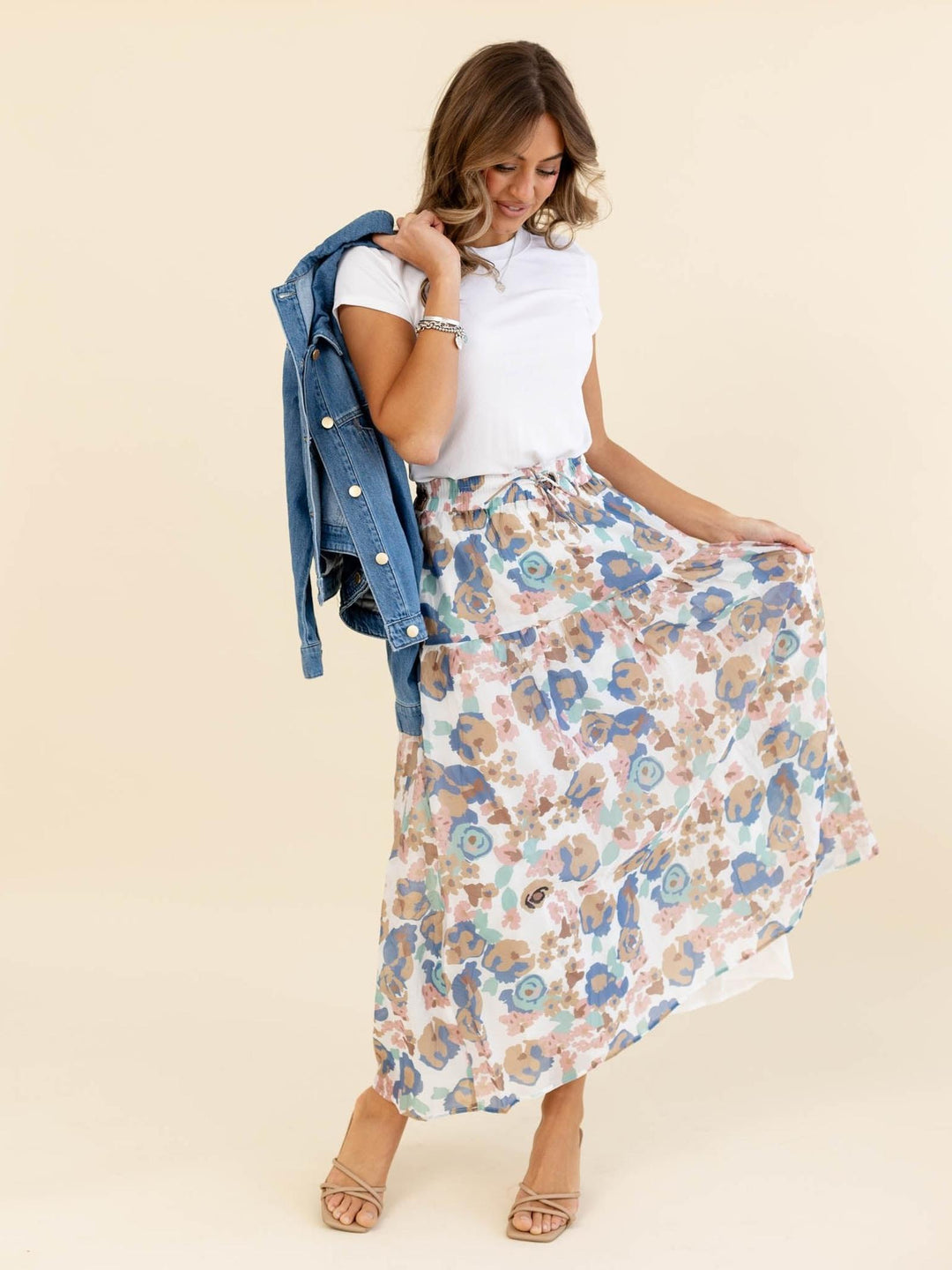 Woman wearing a floral skirt and white top with a denim jacket on a beige background