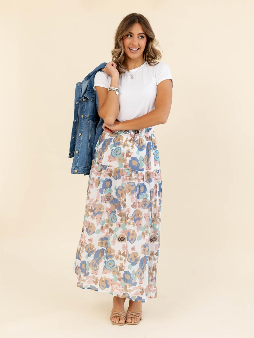 Woman wearing a floral skirt and white top with a denim jacket on a beige background