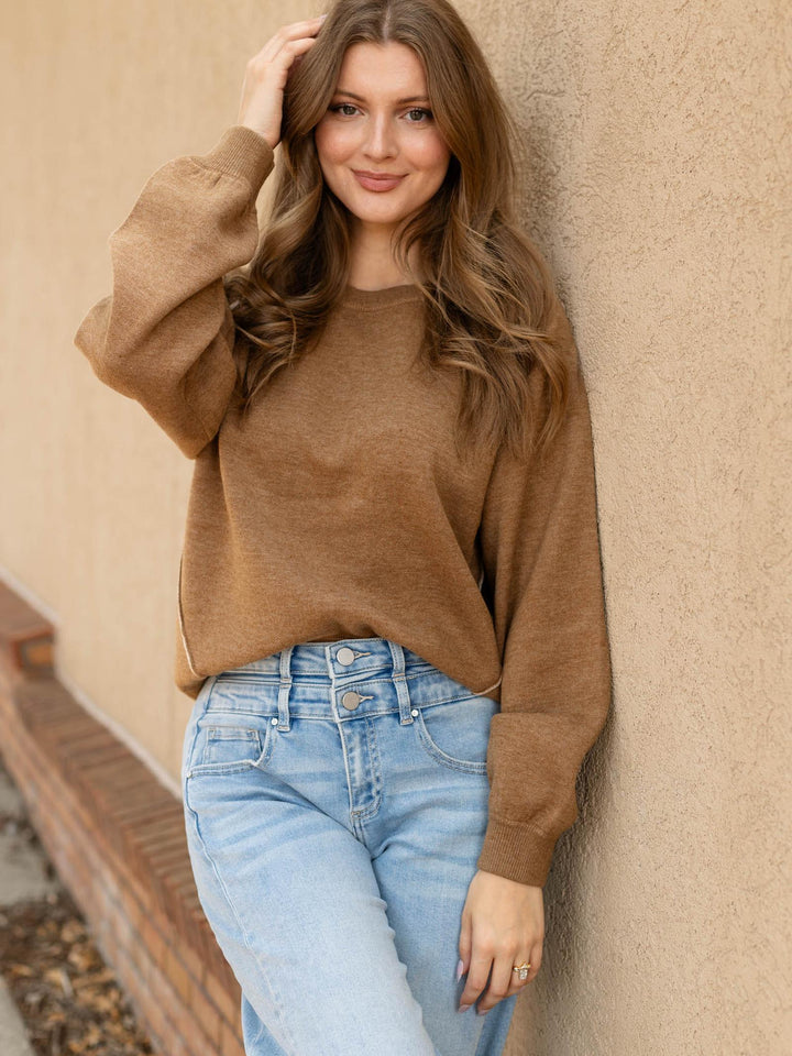 A woman with long wavy brown hair leans against a textured beige wall, wearing a soft camel brown knit crewneck sweater and light wash blue jeans.