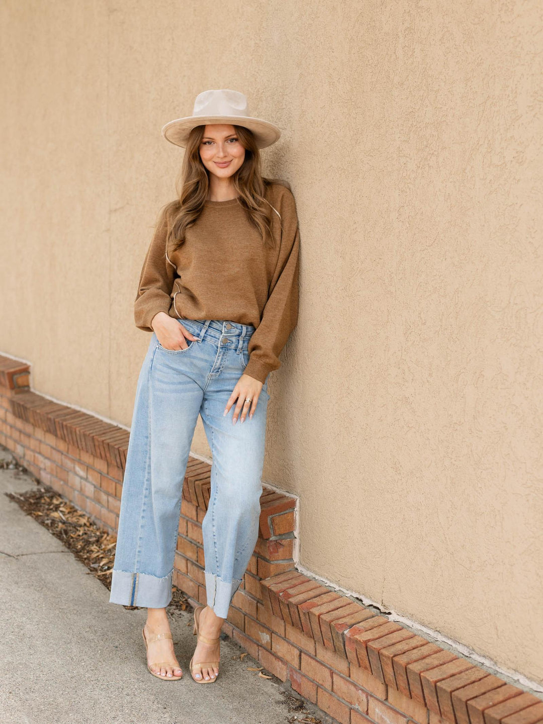 A woman stands outdoors against a beige stucco wall wearing a brown long-sleeve knit top, light blue cropped jeans, a beige wide-brim hat, and nude heels.